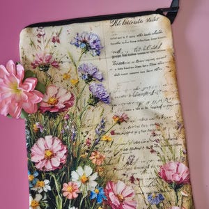 May include: A rectangular floral print pouch with a black zipper and strap. The pouch features a vintage-style design with pink, purple, and white flowers against a backdrop of handwritten script. The text "Nie bettreshe Molls" is visible.
