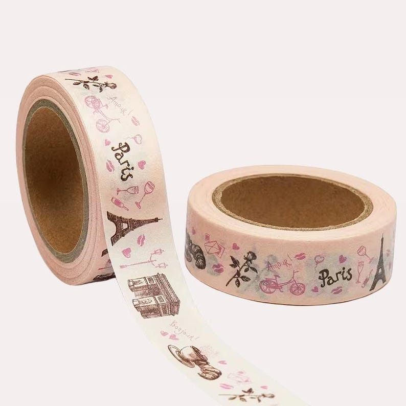 May include: Two rolls of light pink washi tape featuring a repeating pattern of Paris-themed illustrations including the Eiffel Tower, Arc de Triomphe, bicycles, roses, wine glasses, and the text Paris and Amour in a dark brown script font.