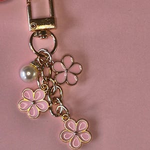 Puede incluir: Un llavero dorado con un cierre de mosquet&oacute;n, adornado con tres colgantes de flores rosas y una perla de imitaci&oacute;n. Los colgantes de flores tienen un contorno blanco y centros dorados. El fondo es de color rosa liso.