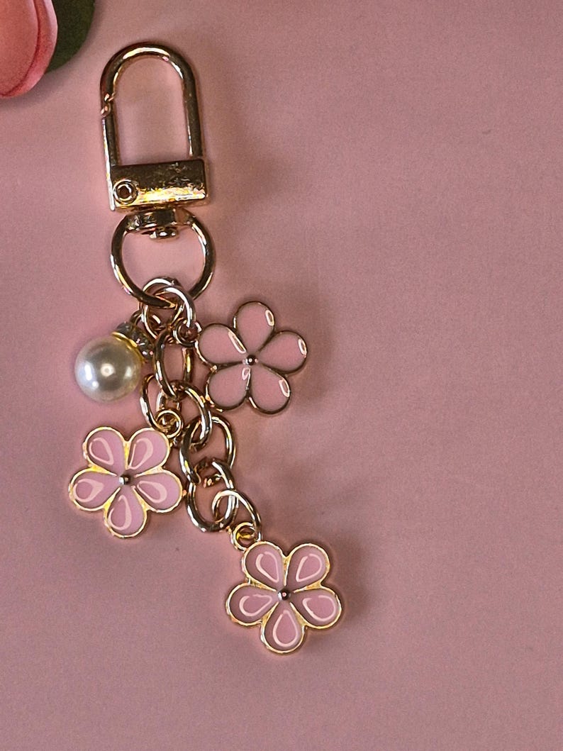 Puede incluir: Un llavero dorado con un cierre de mosquet&oacute;n, adornado con tres colgantes de flores rosas y una perla de imitaci&oacute;n. Los colgantes de flores tienen un contorno blanco y centros dorados. El fondo es de color rosa liso.