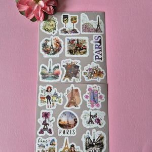 May include: Two sheets of Paris-themed stickers featuring the Eiffel Tower, wine glasses, and floral designs. The stickers are in various shapes and sizes, with the word "Paris" appearing on several. A pink flower is in the upper left corner.