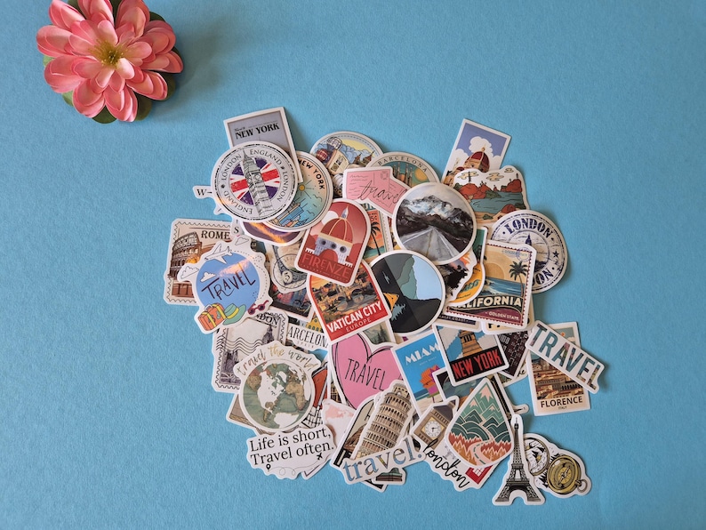 May include: A collection of travel-themed stickers with various designs, including landmarks like the Eiffel Tower and Big Ben, and the text "TRAVEL". The stickers are in various shapes and sizes, with a pink flower in the upper left corner.