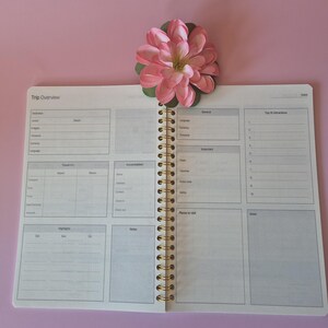 K&ouml;nnte beinhalten: Offener Reiseplaner mit Spiralbindung und goldfarbenen Spiralen. Der Planer ist auf einer Seite mit Abschnitten f&uuml;r Reisedetails, Reiseinformationen und Notizen ge&ouml;ffnet. Eine rosa Blume mit gr&uuml;nen Bl&auml;ttern liegt oben auf dem Planer.