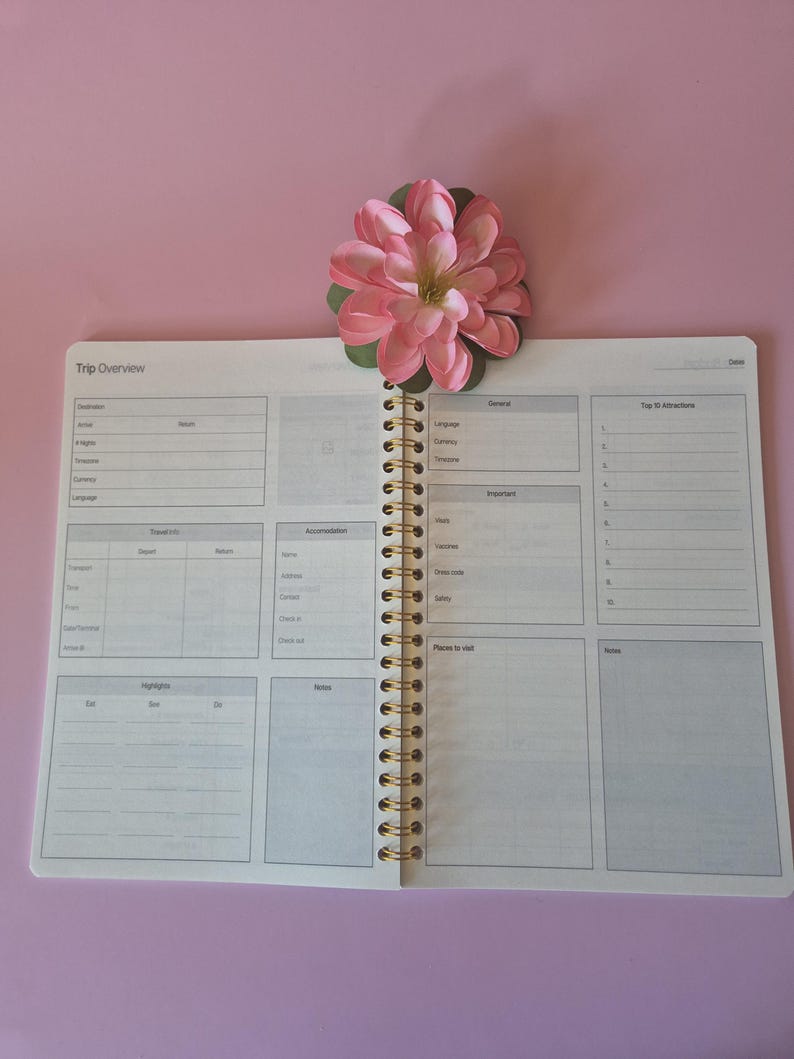 K&ouml;nnte beinhalten: Offener Reiseplaner mit Spiralbindung und goldfarbenen Spiralen. Der Planer ist auf einer Seite mit Abschnitten f&uuml;r Reisedetails, Reiseinformationen und Notizen ge&ouml;ffnet. Eine rosa Blume mit gr&uuml;nen Bl&auml;ttern liegt oben auf dem Planer.