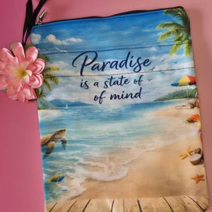 May include: A rectangular beach-themed pouch with a black strap and a pink flower. The pouch features a beach scene with the text "Paradise is a state of mind." The design includes a turtle, palm trees, and a colorful umbrella.