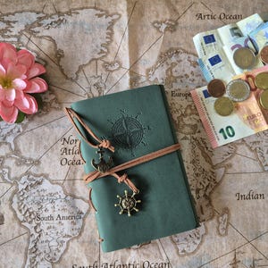 May include: A teal journal with a compass rose design and leather tie, adorned with nautical charms, rests on a vintage world map. A pink flower and Euro currency are also present. The map features ocean names and illustrations of ships.