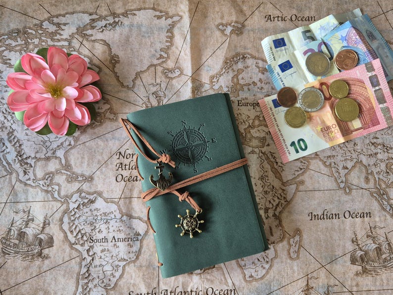 May include: A teal journal with a compass rose design and leather tie, adorned with nautical charms, rests on a vintage world map. A pink flower and Euro currency are also present. The map features ocean names and illustrations of ships.