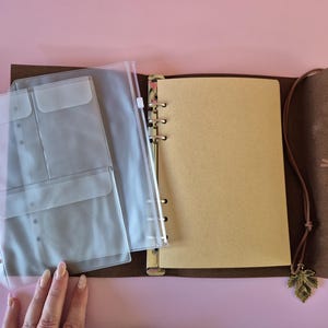 May include: Open brown leather journal with a ring binder, clear plastic pockets, and blank pages. A leather cord with a leaf charm hangs from the journal. The journal is open on a pink surface.
