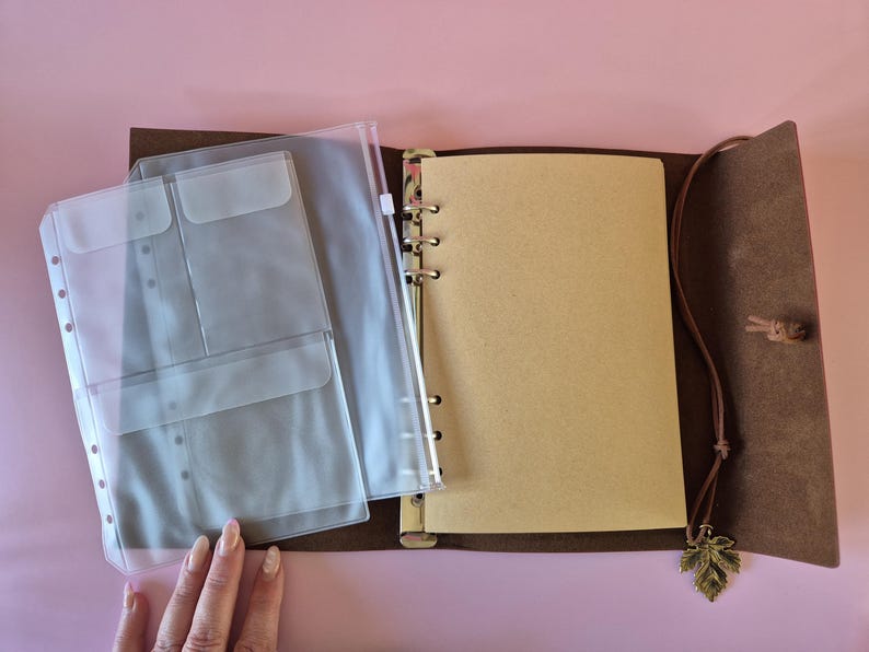May include: Open brown leather journal with a ring binder, clear plastic pockets, and blank pages. A leather cord with a leaf charm hangs from the journal. The journal is open on a pink surface.