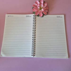 Puede incluir: Un cuaderno abierto, con espiral negra, sobre una superficie rosa. El cuaderno tiene p&aacute;ginas blancas rayadas. Una flor artificial rosa y verde est&aacute; encima. El cuaderno est&aacute; abierto, listo para usar.