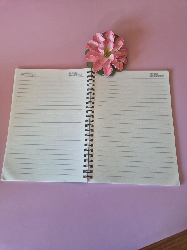 Puede incluir: Un cuaderno abierto, con espiral negra, sobre una superficie rosa. El cuaderno tiene p&aacute;ginas blancas rayadas. Una flor artificial rosa y verde est&aacute; encima. El cuaderno est&aacute; abierto, listo para usar.