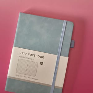 May include: A dusty blue A5 grid notebook with a vertical elastic closure band and a pen loop. A paper belly band features the text GRID NOTEBOOK, High Quality Paper, A5, 100gsm, 14.5cm x 20.3cm, and 160pages, along with a graphic of a grid pattern.