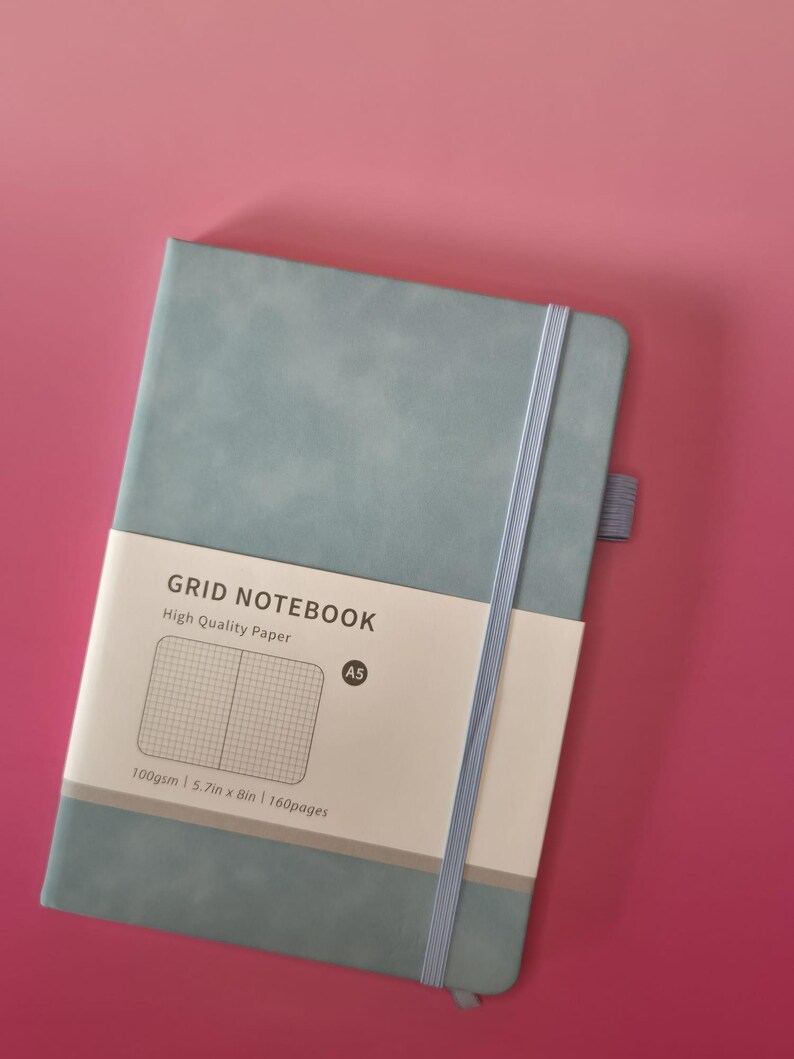 May include: A dusty blue A5 grid notebook with a vertical elastic closure band and a pen loop. A paper belly band features the text GRID NOTEBOOK, High Quality Paper, A5, 100gsm, 14.5cm x 20.3cm, and 160pages, along with a graphic of a grid pattern.