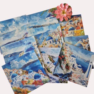 Puede incluir: Una colecci&oacute;n de tarjetas de arte en acuarela con escenas de Santorini, Grecia. Las tarjetas representan edificios blancos con c&uacute;pulas azules, sobre un fondo del mar Egeo y cielos azules. Una flor rosa descansa encima de las tarjetas.