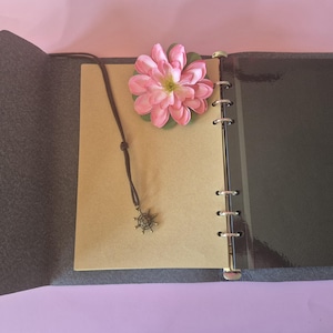 Op de afbeelding: Open grijs vilten tijdschrift met een bruin leren koordketting met een kompasbedel. Een roze bloem rust op de pagina's van het tijdschrift. Het tijdschrift heeft een ringband en een donkergrijze omslag.