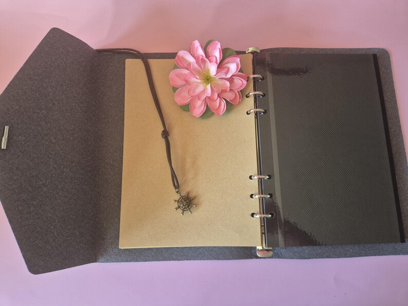 Op de afbeelding: Open grijs vilten tijdschrift met een bruin leren koordketting met een kompasbedel. Een roze bloem rust op de pagina's van het tijdschrift. Het tijdschrift heeft een ringband en een donkergrijze omslag.