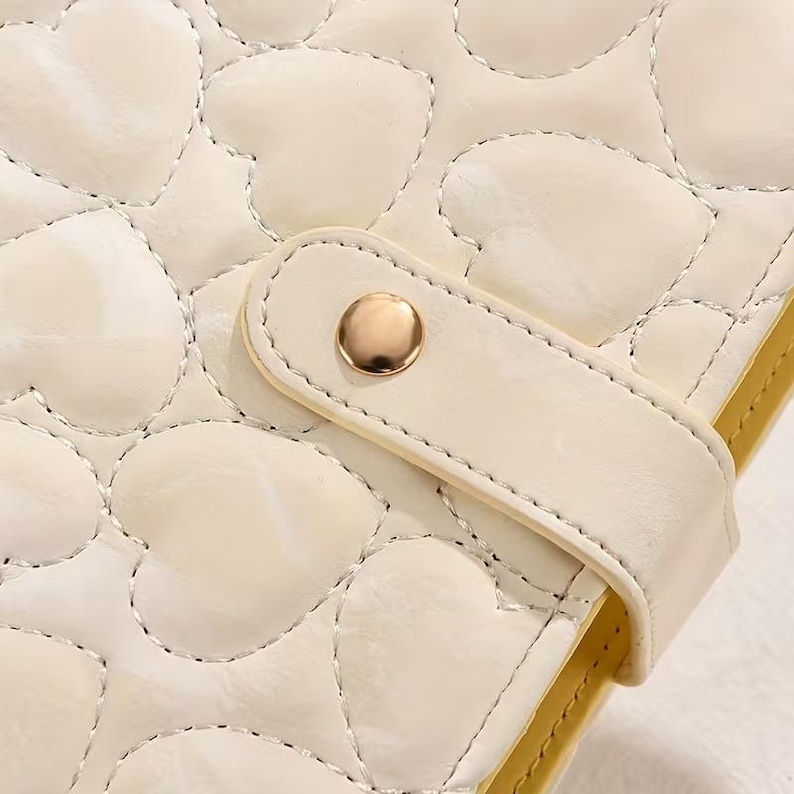 May include: Close-up of a cream-colored, quilted notebook cover with a heart pattern. The cover features a strap with a gold-colored button closure. The edges of the notebook are a pale yellow color.
