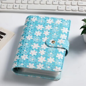 K&ouml;nnte beinhalten: Ein hellblauer Ordner mit floralem Muster und Druckknopfverschluss. Der Ordner hat ein Design mit wei&szlig;en Blumen und Sternen. Der Ordner liegt auf einer wei&szlig;en Oberfl&auml;che in der N&auml;he einer Tastatur.