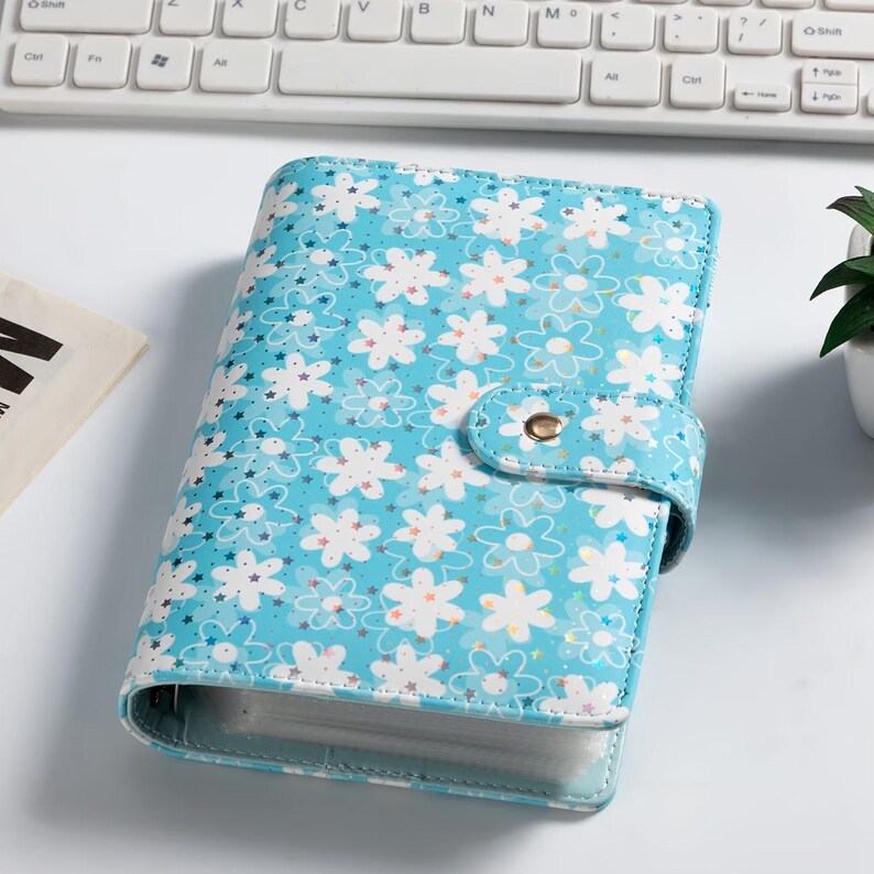 K&ouml;nnte beinhalten: Ein hellblauer Ordner mit floralem Muster und Druckknopfverschluss. Der Ordner hat ein Design mit wei&szlig;en Blumen und Sternen. Der Ordner liegt auf einer wei&szlig;en Oberfl&auml;che in der N&auml;he einer Tastatur.