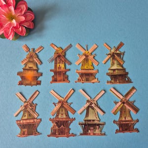 May include: An overhead shot displays eight miniature windmill models with brown, yellow, and green hues, arranged on a blue surface. A pink and red flower is in the upper left corner. The windmills have detailed designs.