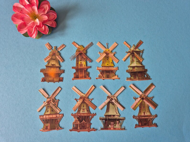 May include: An overhead shot displays eight miniature windmill models with brown, yellow, and green hues, arranged on a blue surface. A pink and red flower is in the upper left corner. The windmills have detailed designs.
