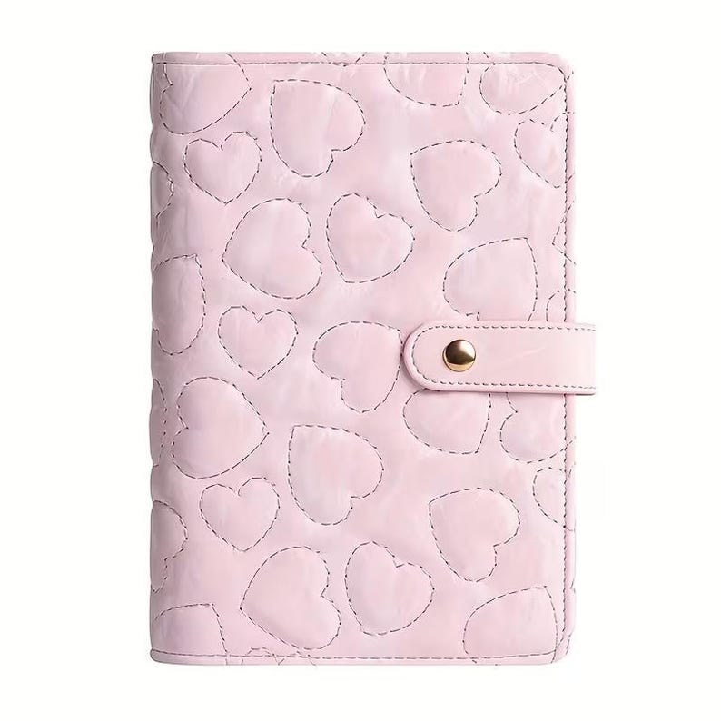 May include: A light pink, quilted planner with a heart pattern. The cover features stitched heart shapes and a matching strap with a gold snap closure. The planner is designed for organization and planning.