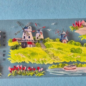 Op de afbeelding: Decoratieve washi tape met een aquarel landschap. Het toont huizen, windmolens en bloemen tegen een lichtblauwe achtergrond. De sc&egrave;ne omvat roodbedekte gebouwen en groene velden, die een dorp uitbeelden.