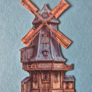 May include: A paper cutout of a windmill with a blue roof and orange blades. The windmill has a wooden exterior with multiple windows and a balcony. The background is a light blue felt.