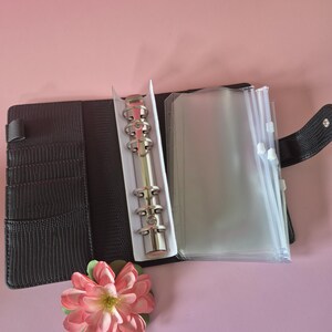Pu&ograve; includere: Raccoglitore in similpelle nera con meccanismo ad anelli in metallo argentato, aperto per rivelare tasche in plastica trasparente. Un fiore artificiale rosa e verde &egrave; posizionato vicino al raccoglitore. Il raccoglitore ha fessure per carte e una chiusura a scatto.