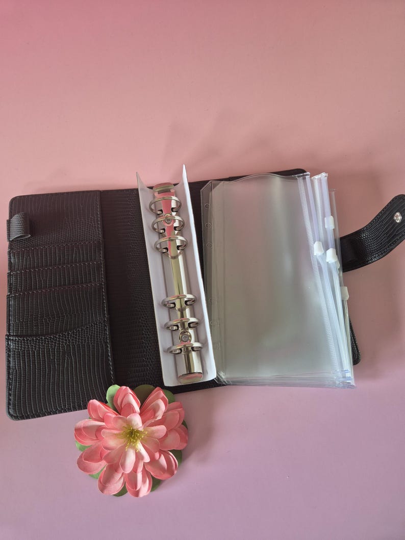 Pu&ograve; includere: Raccoglitore in similpelle nera con meccanismo ad anelli in metallo argentato, aperto per rivelare tasche in plastica trasparente. Un fiore artificiale rosa e verde &egrave; posizionato vicino al raccoglitore. Il raccoglitore ha fessure per carte e una chiusura a scatto.