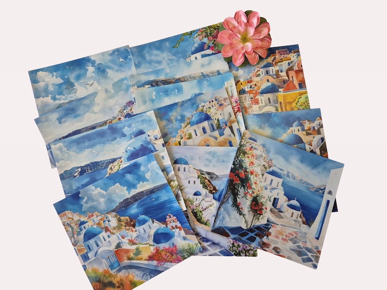 May include: A selection of watercolour art prints showcasing scenes from Santorini, Greece, featuring white buildings, blue domes, and the Aegean Sea. A pink flower sits atop the prints. The prints display various compositions and perspectives.
