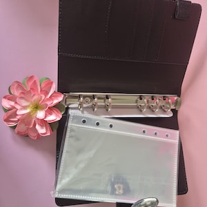 K&ouml;nnte beinhalten: Offener brauner Ordner mit silbernen Ringen und transparenten Kunststofftaschen. Auf der linken Seite befindet sich eine rosa-gr&uuml;ne Blume. Der Ordner ist ge&ouml;ffnet und zeigt Kartenf&auml;cher und einen Druckknopfverschluss mit floralem Design.