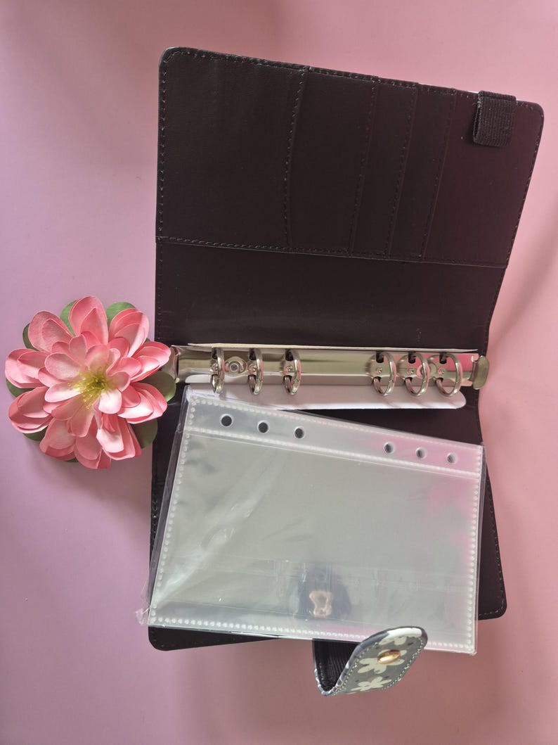 K&ouml;nnte beinhalten: Offener brauner Ordner mit silbernen Ringen und transparenten Kunststofftaschen. Auf der linken Seite befindet sich eine rosa-gr&uuml;ne Blume. Der Ordner ist ge&ouml;ffnet und zeigt Kartenf&auml;cher und einen Druckknopfverschluss mit floralem Design.