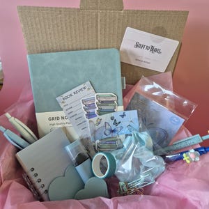 May include: A cardboard box filled with stationery items including a light blue grid notebook, a spiral-bound notepad, a roll of teal tape, a light blue scrunchie, various pens, heart-shaped cutouts, and stickers featuring books and butterflies.