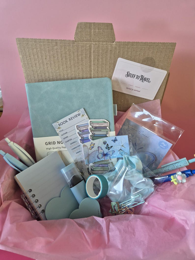 May include: A cardboard box filled with stationery items including a light blue grid notebook, a spiral-bound notepad, a roll of teal tape, a light blue scrunchie, various pens, heart-shaped cutouts, and stickers featuring books and butterflies.