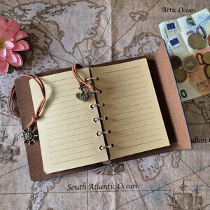 May include: A brown leather-bound notebook with lined pages, open to display the interior. The notebook has a leather cord closure with a ship's wheel charm and a heart charm. A pink flower and Euro currency are also visible.