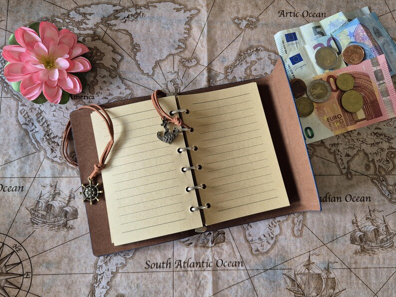 May include: A brown leather-bound notebook with lined pages, open to display the interior. The notebook has a leather cord closure with a ship's wheel charm and a heart charm. A pink flower and Euro currency are also visible.