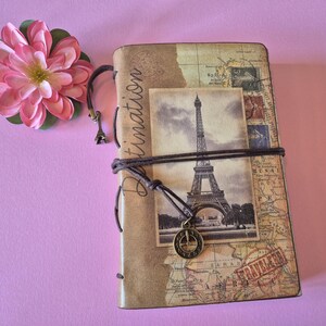 Peut inclure: Un journal de voyage avec un design de carte vintage et une image en noir et blanc de la Tour Eiffel. Le journal est reli&eacute; avec une corde marron et comporte un petit charme de la Tour Eiffel. Une fleur rose repose sur le coin sup&eacute;rieur gauche.