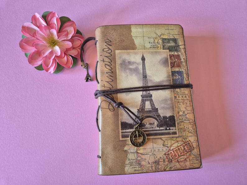 Peut inclure: Un journal de voyage avec un design de carte vintage et une image en noir et blanc de la Tour Eiffel. Le journal est reli&eacute; avec une corde marron et comporte un petit charme de la Tour Eiffel. Une fleur rose repose sur le coin sup&eacute;rieur gauche.