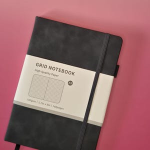 May include: A black A5 grid notebook with a vertical elastic closure band and a paper belly band. The belly band features the text GRID NOTEBOOK, High Quality Paper, A5, 100gsm, 5.7in x 8in, and 160pages, along with a graphic of a grid pattern.