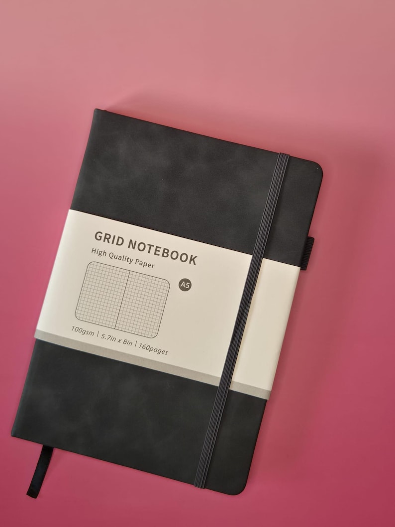May include: A black A5 grid notebook with a vertical elastic closure band and a paper belly band. The belly band features the text GRID NOTEBOOK, High Quality Paper, A5, 100gsm, 5.7in x 8in, and 160pages, along with a graphic of a grid pattern.