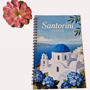 Puede incluir: Cuaderno de espiral con un dise&ntilde;o de Santorini, Grecia. La portada muestra una iglesia con c&uacute;pula azul, edificios blancos e hortensias azules. Una gran flor rosa se coloca cerca del cuaderno.