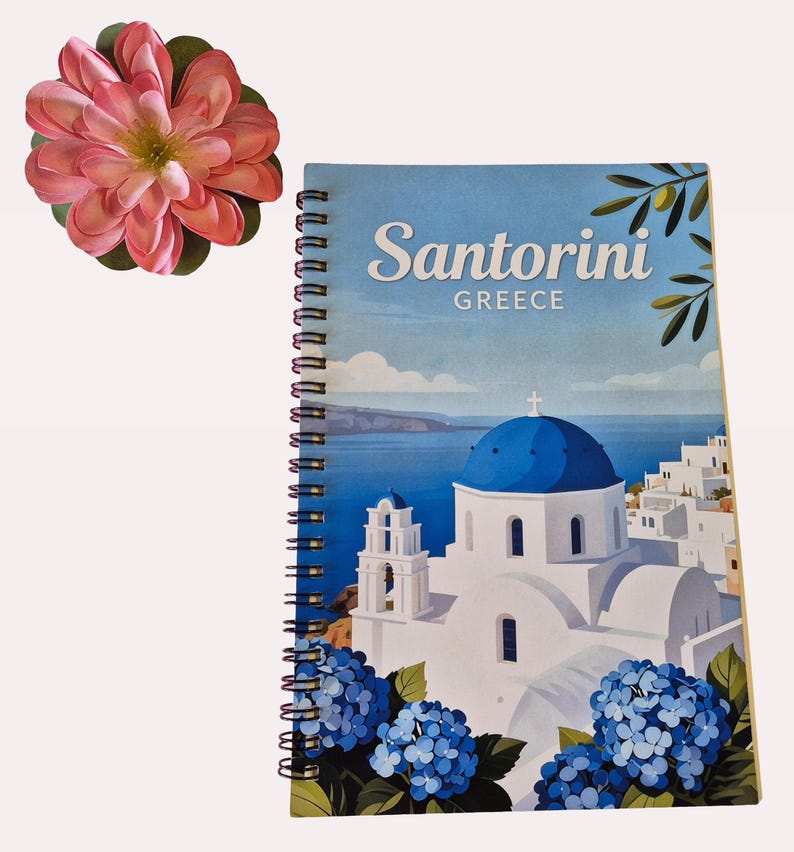 Puede incluir: Cuaderno de espiral con un dise&ntilde;o de Santorini, Grecia. La portada muestra una iglesia con c&uacute;pula azul, edificios blancos e hortensias azules. Una gran flor rosa se coloca cerca del cuaderno.