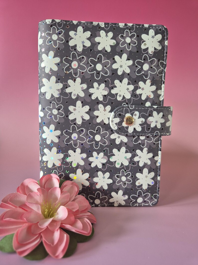 K&ouml;nnte beinhalten: Ein grauer Planer mit floralem Muster und Druckknopfverschluss. Der Planer hat ein sich wiederholendes Muster aus wei&szlig;en Blumen mit schillernden Akzenten. Eine rosa Blume mit gr&uuml;nen Bl&auml;ttern liegt vor dem Planer auf rosa Hintergrund.