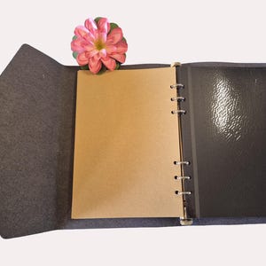 Op de afbeelding: Open grijs vilten tijdschrift met een lichtbruine pagina en een zwarte glanzende pagina. Een roze bloem rust bovenop het tijdschrift. Het tijdschrift is open en onthult de binnenpagina's en de binding.