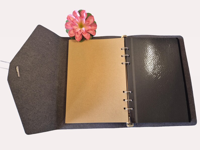 Op de afbeelding: Open grijs vilten tijdschrift met een lichtbruine pagina en een zwarte glanzende pagina. Een roze bloem rust bovenop het tijdschrift. Het tijdschrift is open en onthult de binnenpagina's en de binding.