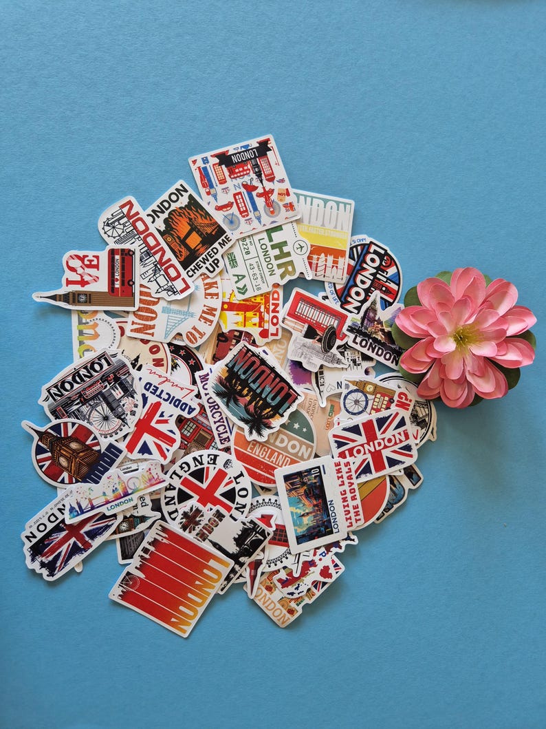 Peut inclure: Un assortiment d'autocollants sur le th&egrave;me de Londres, avec des motifs vari&eacute;s comme l'Union Jack, les cabines t&eacute;l&eacute;phoniques rouges et des paysages urbains. Les autocollants sont dispos&eacute;s sur un fond bleu clair, avec une fleur rose.