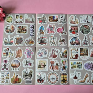 May include: Sheets of stickers with various designs featuring the Eiffel Tower, Parisian scenes, and the word "Paris". The stickers are in various shapes and sizes, with a pink flower in the upper left corner.