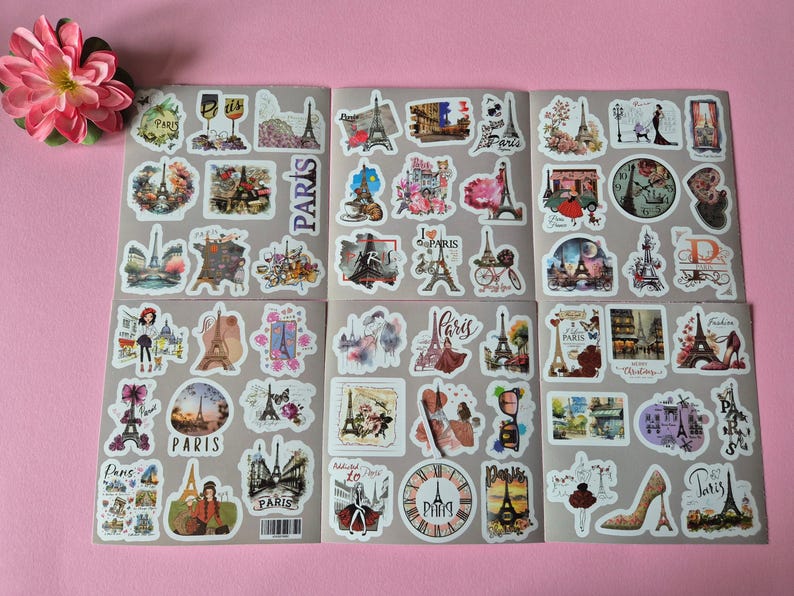 May include: Sheets of stickers with various designs featuring the Eiffel Tower, Parisian scenes, and the word "Paris". The stickers are in various shapes and sizes, with a pink flower in the upper left corner.