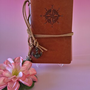 May include: A brown leather journal with a compass rose design embossed on the cover. The journal is tied with a tan leather cord and features a decorative anchor charm. A pink flower with green leaves is in the foreground.